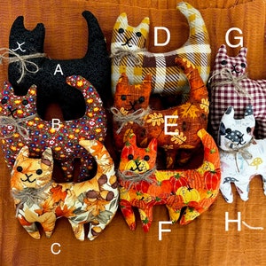 May include: Nine handmade fabric cat toys in various fall-themed patterns. The cats are made of different fabrics, including plaid, gingham, and floral prints. The cats are all about 10 inches tall and have a variety of facial expressions.
