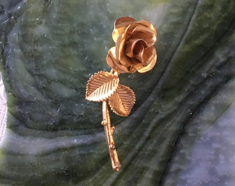 Könnte beinhalten: Eine goldfarbene Rosenbrosche mit einer einzelnen Rose und Blättern. Die Rose ist mit einem Wirbelmuster verziert.
