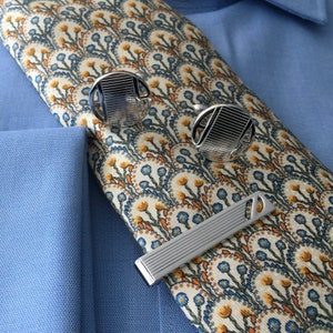 May include: A silver tie bar with a decorative design and two silver cufflinks with a similar design. The cufflinks and tie bar are on a patterned fabric background.