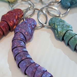 Peut inclure: Un ensemble de porte-clés en forme de dragon enroulés et colorés. Les porte-clés sont faits d'un matériau métallique texturé et sont disponibles dans des tons de violet, turquoise et rouge.