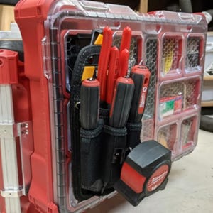 May include: A red toolbox with a clear plastic compartment on the side. The compartment contains a black tool belt with multiple red and black screwdrivers and a black tape measure.