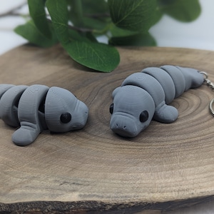Peut inclure: Deux porte-clés en forme de lamantin imprimés en 3D de couleur grise sur une surface en bois. Un lamantin est couché et l'autre est debout. Les deux lamantins ont des yeux noirs et un porte-clés argenté attaché.