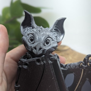 May include: A 3D printed black bat figurine with articulated wings. The bat has a detailed face with large eyes and a textured surface.