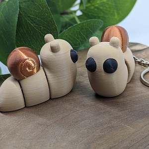 Puede incluir: Dos pequeñas figuritas de caracol impresas en 3D, de color beige, con conchas de color cobre. Un caracol tiene un llavero unido a su concha.