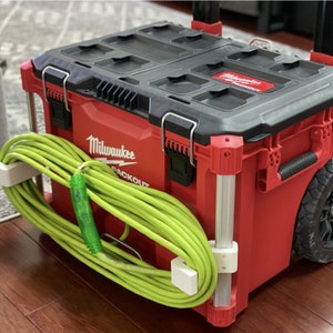 May include: Red Milwaukee Packout rolling toolbox with a black lid and a green extension cord wrapped around the side. The toolbox has a white handle and a black wheel.