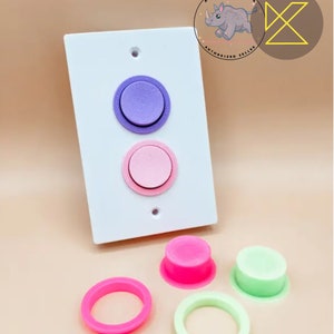 May include: A white light switch plate with two round buttons, one purple and one pink. There are also two smaller round buttons, one pink and one green, and two rings, one pink and one green.