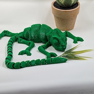 Könnte beinhalten: Ein grüner, 3D-gedruckter Chamäleon mit einem segmentierten Schwanz, auf einer weißen Oberfläche neben einer kleinen Topfpflanze mit Sukkulenten.