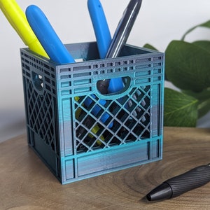 May include: A teal blue plastic milk crate pencil holder with three pens inside. The pens are yellow, blue, and black. The crate is sitting on a wooden surface.