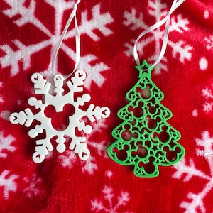 May include: A white snowflake ornament and a green Christmas tree ornament, both featuring cutout Mickey Mouse head silhouettes. Each ornament is crafted from a flat material and includes a thin white ribbon loop for hanging purposes.