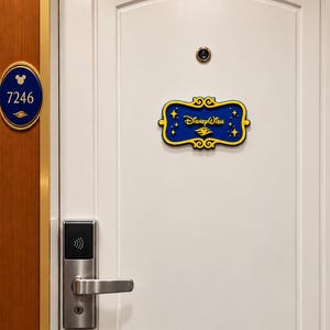 May include: A white door with a silver handle and a digital lock. A blue and yellow sign reads Disney Wish, featuring star accents. A circular peephole is centered above the sign. To the left, a blue oval plaque displays the number 7246.