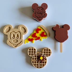May include: A collection of five decorative items including a brown Mickey Mouse head, a tan Mickey Mouse head, a yellow pizza slice with red Mickey-shaped pepperoni, a brown Mickey-shaped popsicle on a stick, and a tan Mickey-shaped waffle.