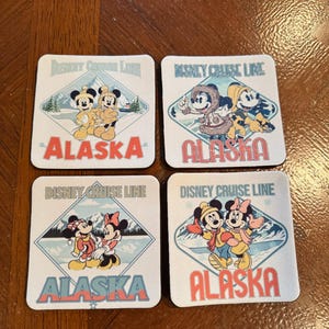 May include: Set of four square coasters featuring Disney characters in winter attire. Each coaster has the text "Disney Cruise Line" and "Alaska" in red and blue. The coasters depict Mickey and Minnie Mouse.
