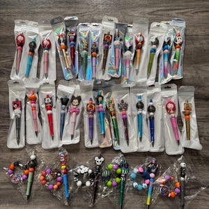 May include: A collection of decorative pens in clear plastic packaging. The pens feature various character-themed toppers in a variety of colors, including red, black, blue, and pink. The pens are displayed on a wooden surface.