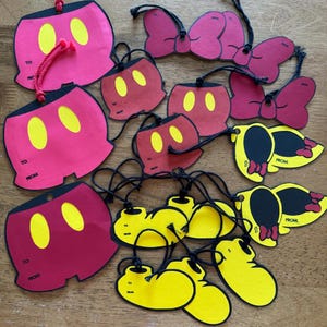 May include: A collection of paper gift tags featuring red shorts with yellow ovals, pink bows, yellow shoes, and yellow silhouettes with pink bows. Each tag includes black string and printed text that reads TO: and FROM: for labeling.