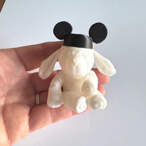 May include: A small white figurine of a seated dog with long floppy ears. The dog is wearing a black cap with two round black ears attached to the top, resembling a classic mouse-ear hat. The figure has a smooth, matte finish.