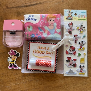 May include: A collection of items including a pink watermelon scented hand sanitizer, a pack of Kleenex tissues with Cinderella, a Minnie Mouse sticker, a lip balm labeled Have a Good Day, a white Mickey Mouse shaped clip, and a sheet of Disney stickers.