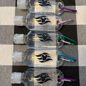 May include: Five clear plastic hand sanitizer bottles with a black Mickey Mouse head and three waves logo. Each bottle has a silver carabiner attached.
