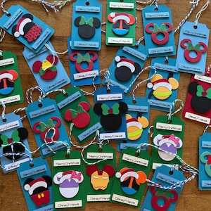 May include: A collection of rectangular paper gift tags in blue, green, and red, each featuring a die-cut character head or ornament shape, a white label with the text Merry Christmas, and a secondary label with the text Disney Friends.