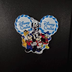 May include: A Disney Cruise magnet in the shape of Mickey Mouse's head. The magnet features Mickey, Minnie, Donald, Goofy, and Pluto. The words "Disney Cruise" are written in blue on a white background.