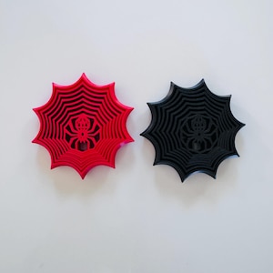 May include: Two circular coasters featuring a spider and web design. One coaster is bright red and the other is solid black. Both items have a raised, multi-layered web pattern radiating from a central spider figure with eight legs.
