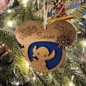 May include: Wooden Disney-themed Christmas ornament in the shape of Mickey Mouse ears. The ornament features the text "Disney Destiny Sarah 2025" and a laser-cut image of Stitch. A snowflake is also included.