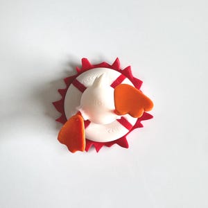 May include: A 3D printed figurine of a white duck with orange feet, positioned inside a white life preserver ring with red stripes. The entire assembly is mounted on a red, jagged, star-shaped base, viewed from directly above.