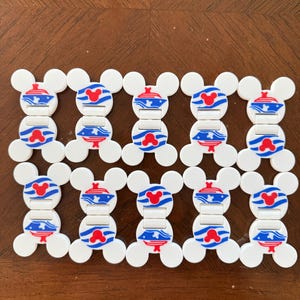May include: Ten white plastic clips shaped like mouse heads, arranged in two rows of five. Each clip features a central graphic of a blue cruise ship with a red mouse head icon above it, set against blue wavy lines on a white background.