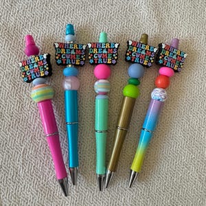 May include: Five pens with colorful beads and a black topper featuring the text WHERE DREAMS COME TRUE in multi-colored letters. The pens have metallic barrels in shades of pink, blue, mint green, gold, and a blue-to-yellow gradient finish.