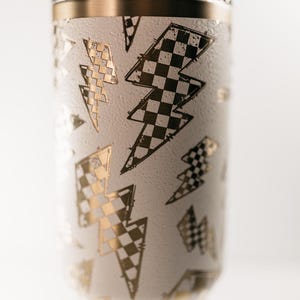 Op de afbeelding: Witte beker met gouden en zwarte bliksemontwerpen. De beker heeft een gouden rand en een doorzichtig deksel. De bliksems zijn in een dambordpatroon. Een doorzichtig rietje is zichtbaar.