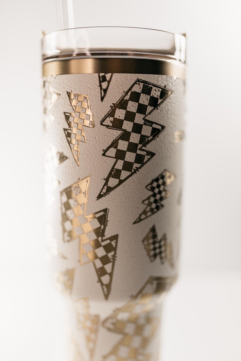 Op de afbeelding: Witte beker met gouden en zwarte bliksemontwerpen. De beker heeft een gouden rand en een doorzichtig deksel. De bliksems zijn in een dambordpatroon. Een doorzichtig rietje is zichtbaar.