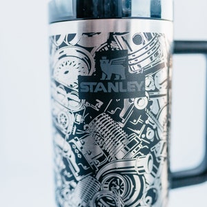 May include: Stainless steel travel mug with a black handle and lid. The mug features a detailed, etched design of engine components and the Stanley logo in black. The mug is designed to keep beverages hot or cold.