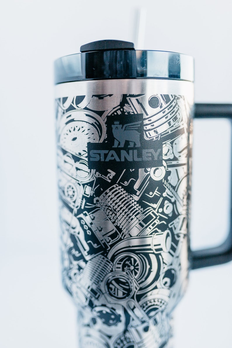 May include: Stainless steel travel mug with a black handle and lid. The mug features a detailed, etched design of engine components and the Stanley logo in black. The mug is designed to keep beverages hot or cold.