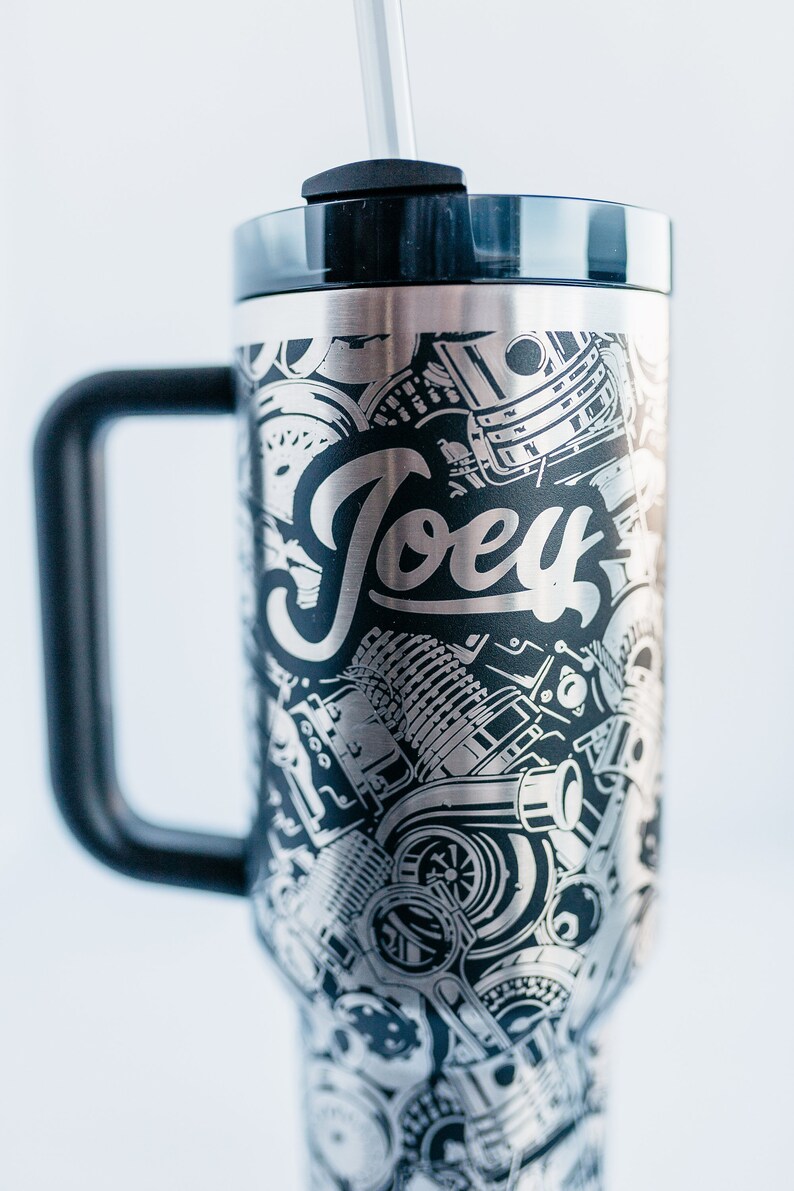 May include: Stainless steel travel mug with a black handle and lid. The mug features a detailed engraved design of engine parts and the name "Joey" in a stylized font. Includes a black straw.
