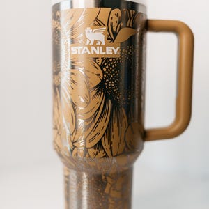 Pu&ograve; includere: Un grande bicchiere da viaggio Stanley con un design a girasole nei toni del marrone, nero e argento. Il bicchiere ha un manico e un coperchio bianco. Il logo Stanley &egrave; visibile sul davanti.