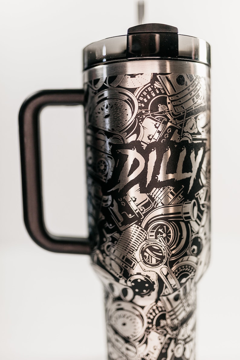 May include: Stainless steel travel mug with a black handle and lid. The mug is covered in a detailed, etched design of engine parts and the word "DILLY" in a bold font. The mug is designed to keep beverages hot or cold.