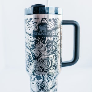 May include: A stainless steel Stanley tumbler with a black handle and lid. The tumbler is covered in a detailed, etched pattern of engine parts and machinery. The Stanley logo is prominently displayed. A clear straw is inserted into the lid.