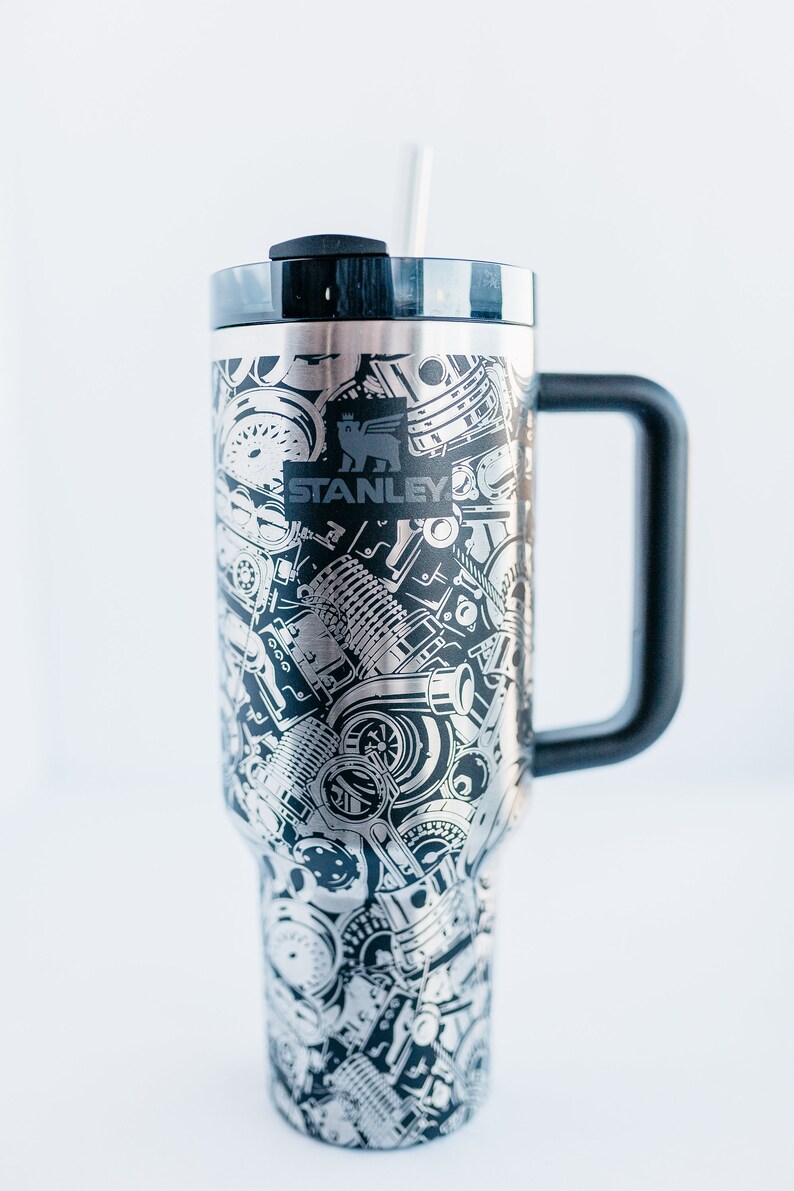 May include: A stainless steel Stanley tumbler with a black handle and lid. The tumbler is covered in a detailed, etched pattern of engine parts and machinery. The Stanley logo is prominently displayed. A clear straw is inserted into the lid.