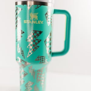 Op de afbeelding: Een turquoise Stanley-beker met een handvat en een deksel. Het heeft een patroon van zilveren en zwart geruite bliksemschichten. Het Stanley-logo is groen gedrukt.