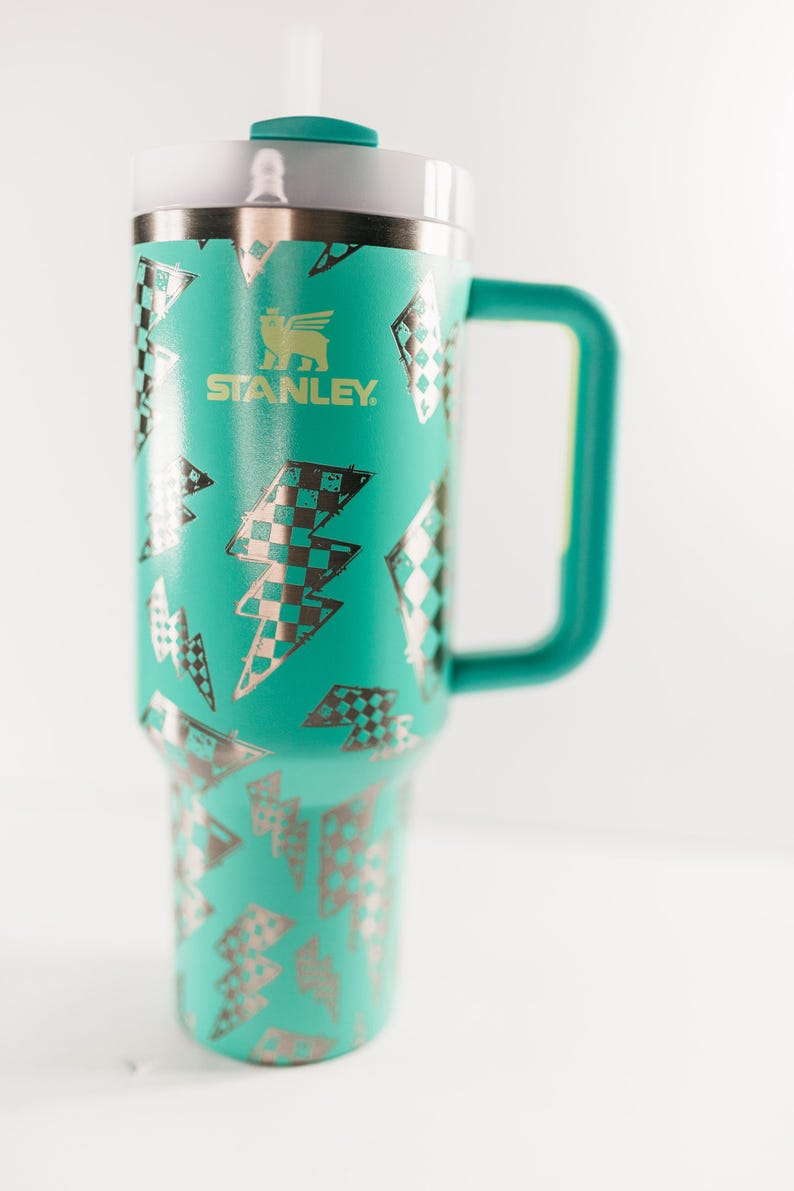 Op de afbeelding: Een turquoise Stanley-beker met een handvat en een deksel. Het heeft een patroon van zilveren en zwart geruite bliksemschichten. Het Stanley-logo is groen gedrukt.
