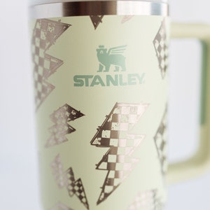 Op de afbeelding: Een lichtgroene Stanley reismok met een handvat en deksel. De mok heeft een herhalend patroon van bliksemschichten met een dambordontwerp. Het Stanley-logo is groen gedrukt.