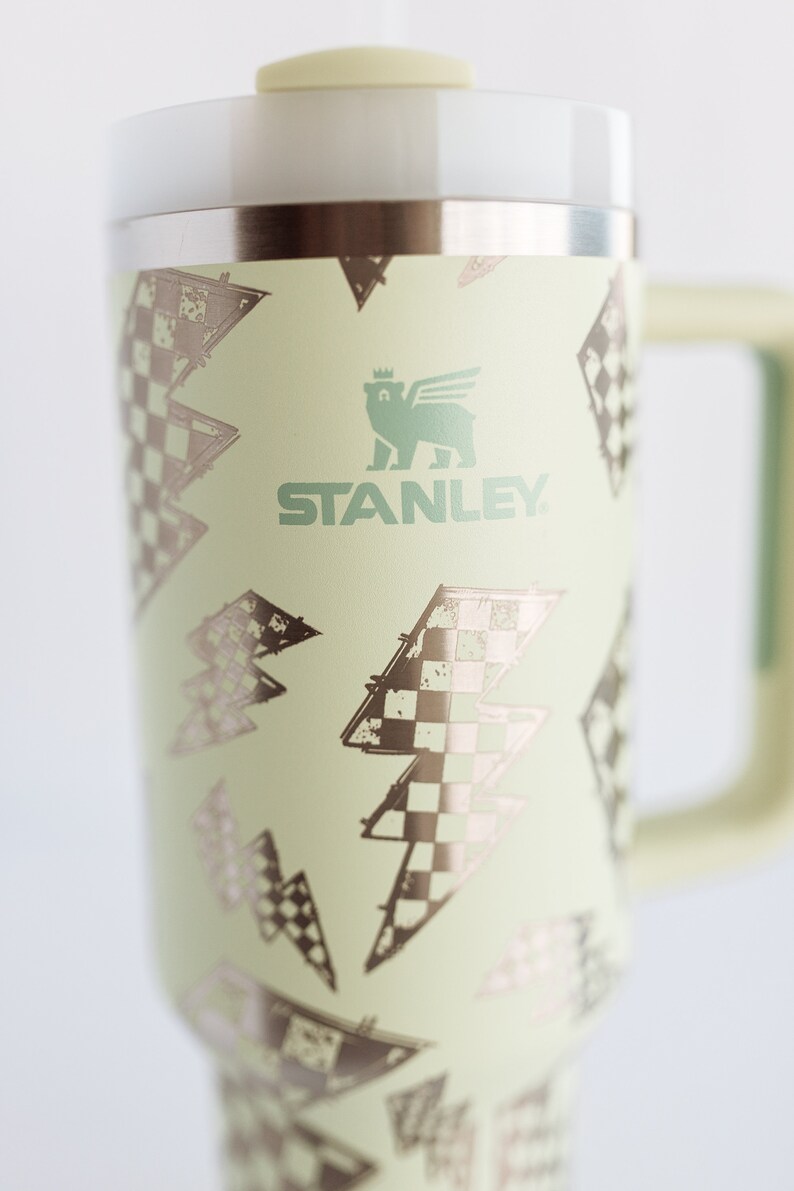 Op de afbeelding: Een lichtgroene Stanley reismok met een handvat en deksel. De mok heeft een herhalend patroon van bliksemschichten met een dambordontwerp. Het Stanley-logo is groen gedrukt.