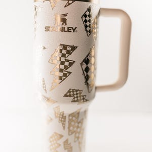 Op de afbeelding: Witte en gouden Stanley-beker met een handvat, met een herhalend patroon van gouden bliksemschichten en dambordontwerpen. De beker heeft een beige handvat, deksel en rietje. Het Stanley-logo is zichtbaar.