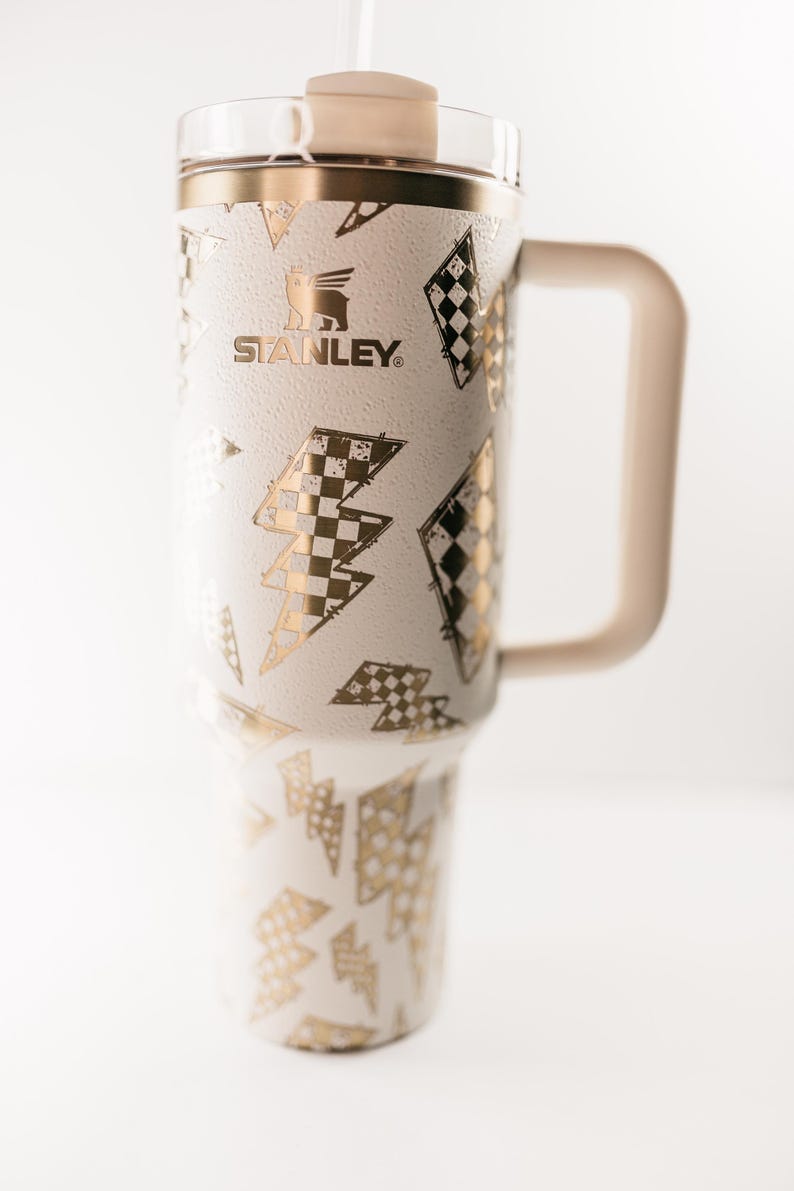 Op de afbeelding: Witte en gouden Stanley-beker met een handvat, met een herhalend patroon van gouden bliksemschichten en dambordontwerpen. De beker heeft een beige handvat, deksel en rietje. Het Stanley-logo is zichtbaar.