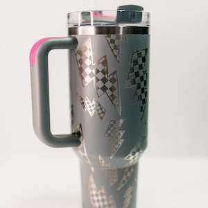 Op de afbeelding: Grijze ge&iuml;soleerde beker met een doorzichtig deksel en een grijze handgreep met een roze accent. De beker heeft een patroon van zilveren bliksemschichten met een geblokt vlagontwerp. Een doorzichtig rietje is in het deksel gestoken.