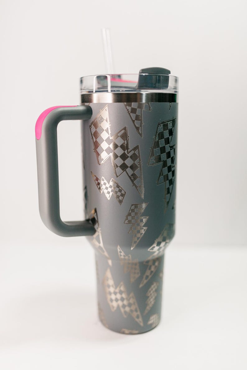 Op de afbeelding: Grijze ge&iuml;soleerde beker met een doorzichtig deksel en een grijze handgreep met een roze accent. De beker heeft een patroon van zilveren bliksemschichten met een geblokt vlagontwerp. Een doorzichtig rietje is in het deksel gestoken.