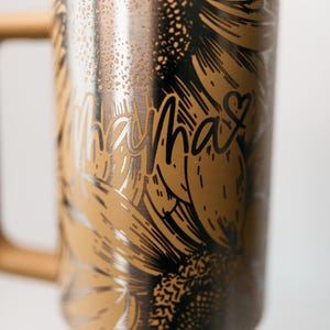 Pu&ograve; includere: Una tazza da viaggio in acciaio inossidabile con coperchio bianco e manico in legno. La tazza presenta un design di girasole dorato con la parola "Mama" in corsivo. Un cuore &egrave; accanto alla parola "Mama".