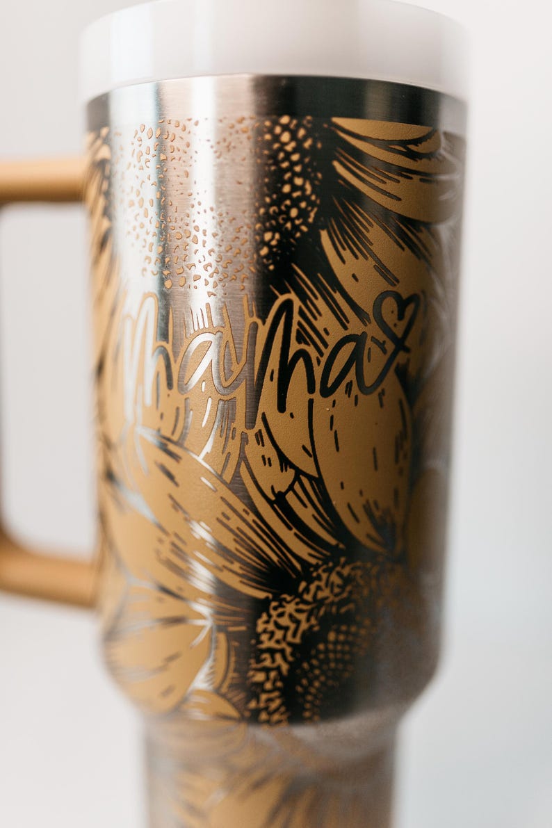 Pu&ograve; includere: Una tazza da viaggio in acciaio inossidabile con coperchio bianco e manico in legno. La tazza presenta un design di girasole dorato con la parola "Mama" in corsivo. Un cuore &egrave; accanto alla parola "Mama".