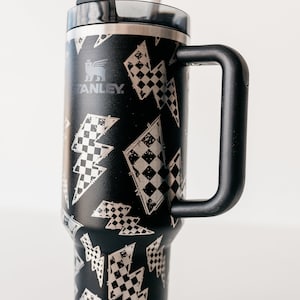 Op de afbeelding: Zwarte Stanley beker met een handvat, versierd met zilveren bliksemschichten en een geblokt vlagpatroon. Het heeft een zwart deksel en een zilveren rietje. Het Stanley-logo is zichtbaar.