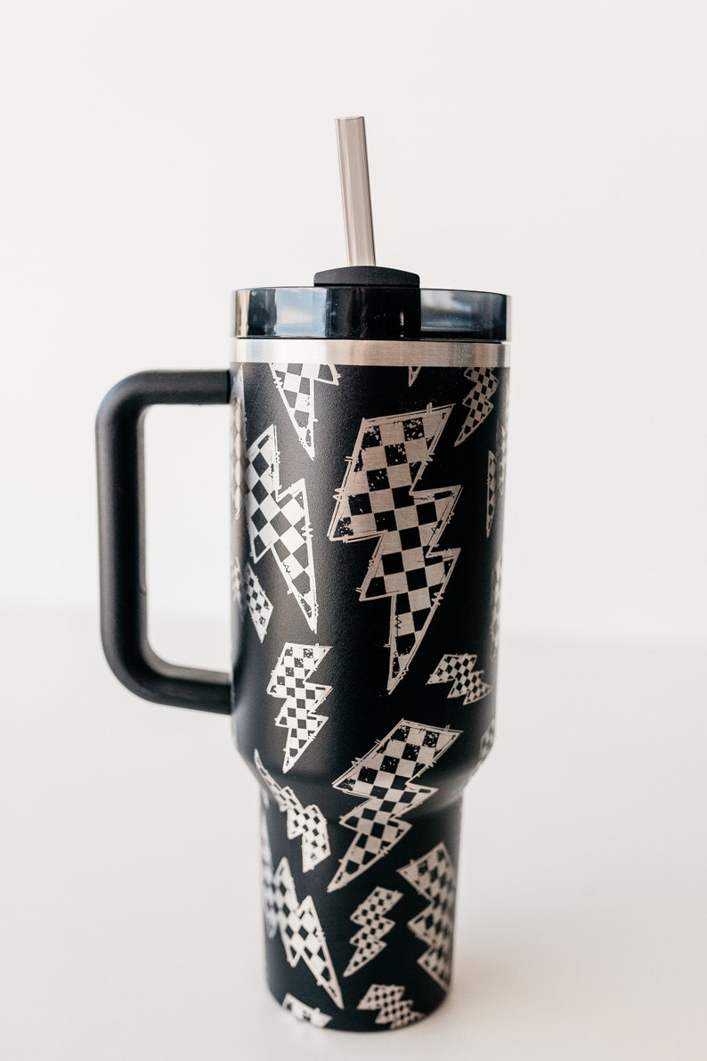 Op de afbeelding: Zwarte ge&iuml;soleerde beker met een handvat en een deksel, met een zilveren rietje. De beker is versierd met een patroon van bliksemschichten gevuld met een dambordontwerp. De handgreep en het deksel zijn zwart.