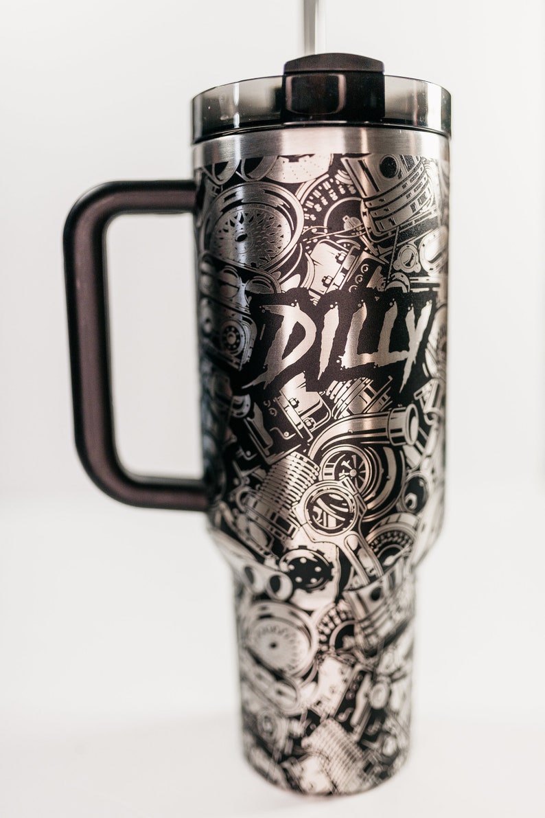 May include: Stainless steel travel mug with a black handle and lid. The mug is covered in a detailed black and silver design of engine parts and the word "DILLY".